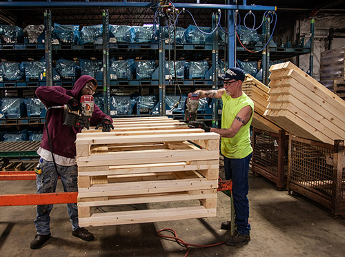 one man wearing maroon hoodie and jeans and another man wearing neon yellow cut-off t-shirt and jeans drill into a custom wooden pallet