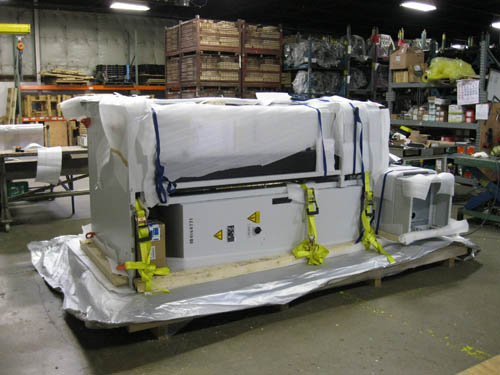 A large machine is secured to a flat wooden pallet with yellow buckle ties. There is styrofoam wrapping on top of the machine that is secured with black rope. 